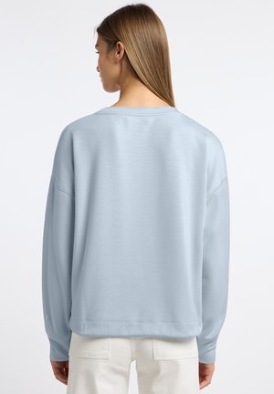 Vrouw met steil lichtbruin haar draagt een lichtblauwe trui met lange mouwen en een witte broek, gezien van achteren tegen een witte achtergrond.