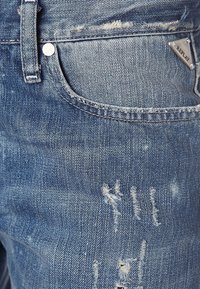 Světle modré džínové kalhoty s opotřebeným vzhledem, viditelnými vzory opotřebení, malou stříbrnou nášivkou a klasickým designem pěti kapes.