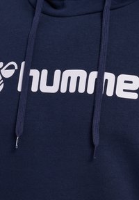 Navy-Hoodie aus weichem Material, mit einem großen weißen "humme"-Logo und Zugbändern für die Kapuze. Texturierte Oberfläche mit minimalen Details.