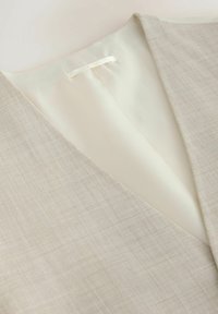 Gros plan sur le col d'un blazer beige texturé montrant une doublure satinée crème lisse et une boucle interne pour suspendre.