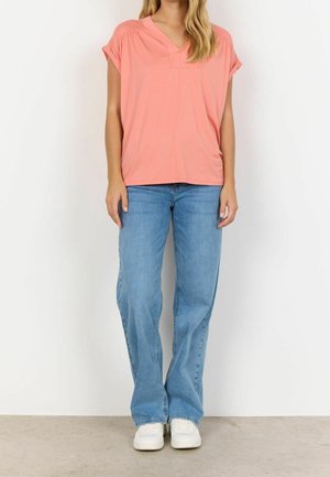 Femme portant un haut corail ample à manches courtes, un jean bleu clair à jambes larges et des baskets blanches, debout devant un fond blanc uni.