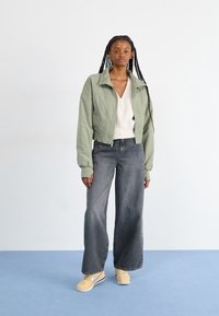 Kvinna med långa flätor som står i en ljusgrön zipjacka, vit V-ringad topp, vida svarta jeans med blekta detaljer och beigefärgade Nike-sneakers på en blå golv.