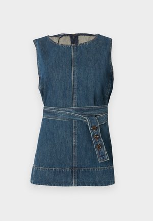 Vrouw met een mouwloos denim top met ceintuur, wijd uitlopende witte broek en zwarte puntige schoenen, staand met de hand op haar heup.