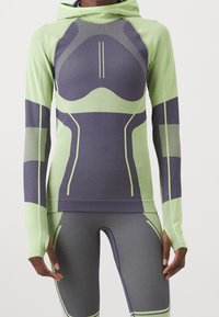 Närbild av en person i en åtsittande sportoutfit med ljusgröna och mörkgrå geometriska mönster och tumhål på ärmarna.