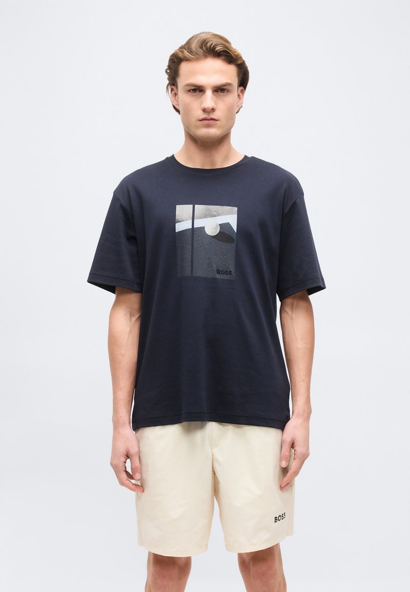 Νέος άνδρας που στέκεται φορώντας μαύρο γραφικό μπλουζάκι και εκρού σορτς με λογότυπο BOSS στο αριστερό πόδι, σε απλό φόντο.