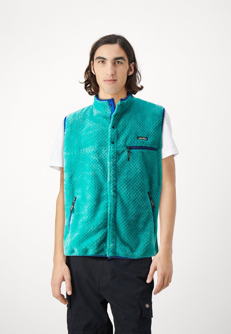 Turkis fleece vest med diamantstruktur, glidelåslommer i siden, ståkrage og kontrasterende blå kant. Logo vist på brystet.