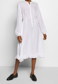 Biała, długa sukienka z długim rękawem, wykonana z przezroczystego materiału, z guzikami z przodu, ściągaczem w talii i falbaną na dole. W połączeniu z czarnymi płaskimi butami.