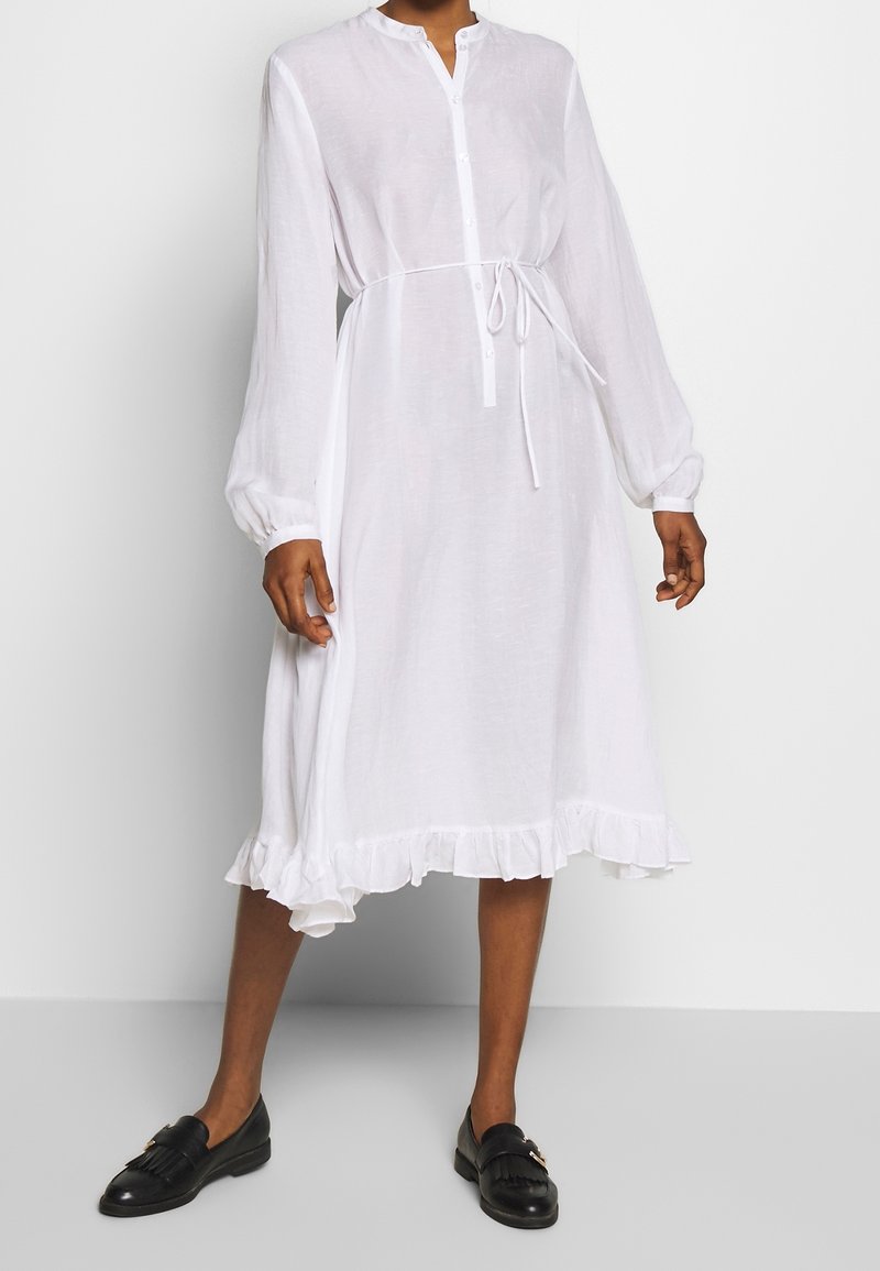 Biała, długa sukienka z długim rękawem, wykonana z przezroczystego materiału, z guzikami z przodu, ściągaczem w talii i falbaną na dole. W połączeniu z czarnymi płaskimi butami.