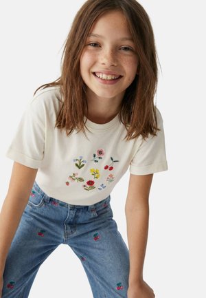 Fille souriante aux cheveux bruns portant un t-shirt crème avec des broderies florales et de cerises, ainsi qu'un jean bleu avec des motifs de cerises, sur un fond blanc.