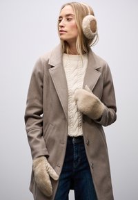 Beżowy wełniany płaszcz z kołnierzem z klapami, zestawiony z kremowym swetrem w warkocze, ciemnymi dżinsami oraz miękkimi beżowymi nausznikami i rękawiczkami.