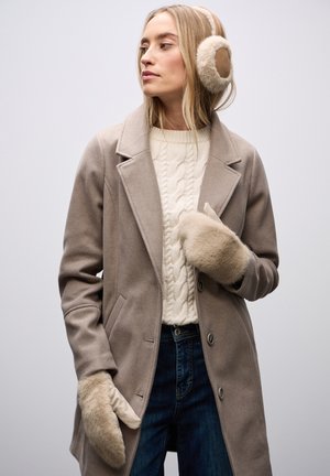 Béžový vlněný kabát s klopovým límcem, doplněný o smetanový svetr s copánkovým vzorem, tmavé džíny a měkké hnědé chrániče uší a rukavice.