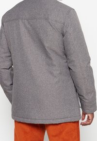 Cappotto grigio strutturato con un design testurizzato e maniche lunghe, caratterizzato da dettagli delle cuciture e un taglio dritto, abbinato a pantaloni arancioni.