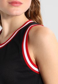 Mustat urheilulliset hihaton paita, jossa on verkko-tekstuuri, punaiset ja valkoiset raidat kauluksessa ja käsivarsien aukkoissa, korostaen urheilullista muotoilua.