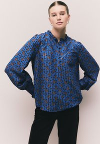 Niebieska bluzka w wzory z motywem kwiatowym, plisowaną fakturą, wysokim kołnierzem i długimi, bufkami rękawów. Z ozdobnym przeszyciem wokół dekoltu.