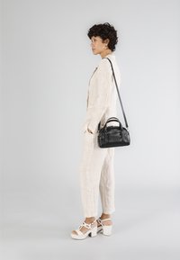 Zwarte leren handtas met korte hengsels en een schouderband, gedragen door een persoon in een lichtbeige, gestructureerde jumpsuit en witte blokhakken.
