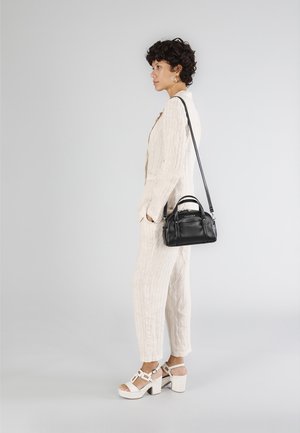 Borsa a tracolla in pelle nera con manici corti, indossata da una persona in una tuta testurizzata beige chiaro e sandali bianchi con tacco quadra.