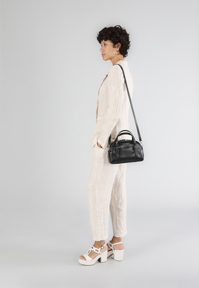 Zwarte leren handtas met korte hengsels en een schouderband, gedragen door een persoon in een lichtbeige, gestructureerde jumpsuit en witte blokhakken.