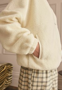 Cream-colored fleece jacket with a textured, fluffy surface, featuring a side pocket and ribbed cuffs, paired with plaid pants.