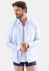 Chemise bleu clair à manches longues, en coton, au design légèrement ajusté. Portée sur un t-shirt à col en V blanc et un short bleu.