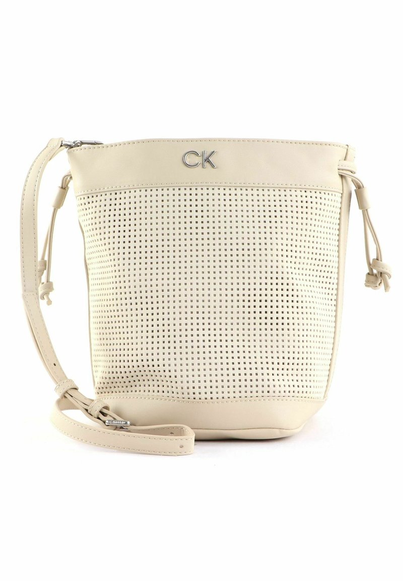 Sac bandoulière en cuir beige avec devant perforé, sangle réglable, fermeture éclair et logo métallique "CK" au centre supérieur.