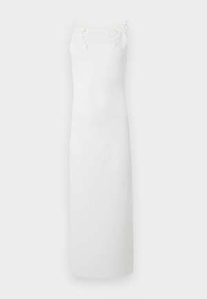 Femeie care poartă o rochie midi albă fără mâneci cu bretele florale, asortată cu pantofi albi cu vârf ascuțit și o brățară de gleznă cu perle, stând pe un fundal simplu.