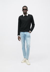 Jersey negro de manga larga con cuello blanco, jeans azul claro y zapatillas crema; gafas de sol completan el conjunto. Diseño sencillo, ajuste casual.
