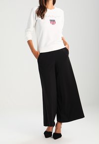 Mujer vistiendo un sudadera blanca con un escudo bordado, pantalones de pierna ancha negros y tacones de punta negra, de pie con las manos en los bolsillos.