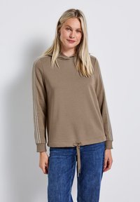 Felpa marrone con coulisse sull'orlo, caratterizzata da strisce verticali sulle maniche, realizzata in tessuto morbido. Indossata con jeans di denim blu.