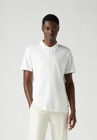 Vit polo skjorta med krage, med tre knappar. Subtil röd brodyr på bröstet, tillverkad av ett mjukt, strukturerat tyg.