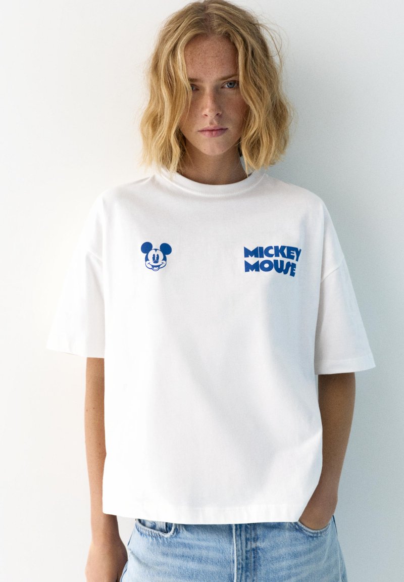 Jeune femme aux cheveux blonds mi-longs portant un T-shirt blanc oversize avec un graphique bleu de Mickey Mouse et du texte, associé à un jean bleu clair.