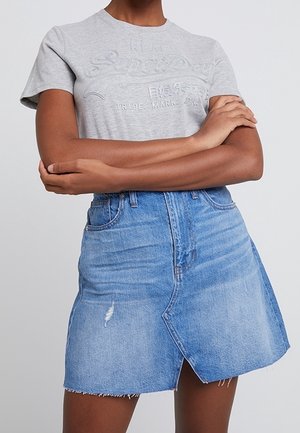 Person iført en lysegrå Superdry t-shirt og en blå denim nederdel med en frontslids og frynset kant, med armene krydsede.