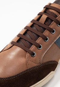 Baskets en cuir et suède marron avec des lacets marron foncé, des œillets argentés et un accent bleu. Présente une surface lisse et texturée.