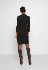 Robe noire ajustée avec des manches trois-quarts, un col haut et une coupe droite, associée à des bottes marron texturées hauteur genoux.