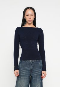 Tmavomodrý rebrovaný pletený sveter so širokým výstrihom, priliehavým živôtikom a dlhými rukávmi zdobenými gombíkmi na manžetách. Nosený s džínsami so širokými nohavicami.