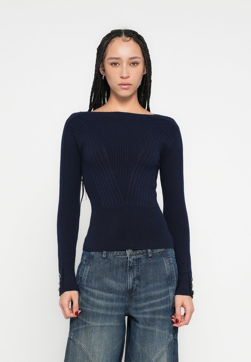 Tmavomodrý rebrovaný pletený sveter so širokým výstrihom, priliehavým živôtikom a dlhými rukávmi zdobenými gombíkmi na manžetách. Nosený s džínsami so širokými nohavicami.