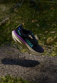 Chaussure de sport noire avec une semelle intermédiaire crème claire, accents violets et teal, présentant un design texturé et une marque visible sur le côté.