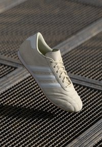 Šviesiai bežiniai futbolo sportbačiai su tekstūruota odine viršutine dalimi, baltais odiniais dryžiais ir guminiais padais, turintys žemą profilį ir apvalią noselę.