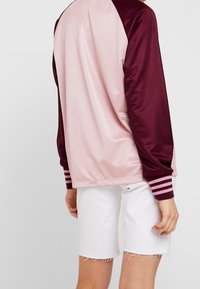 Långärmad sporttröja med rosa kropp och vinröda ärmar. Har randiga detaljer vid ärmsluten. Bärs tillsammans med vita fransade shorts.