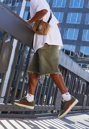 Homme portant des chaussettes blanches Puma, des baskets Puma vertes et rouges, un short kaki et une chemise blanche, debout sur la rambarde métallique d'un pont en milieu urbain.