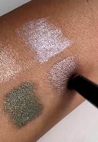 Prøver av tre teksturerte øyenskygger på huden: en glitrende lys rosa, en metallisk rosé og en glitrende olivengrønn, påført med en kost.