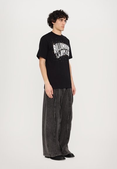 T-shirt graphique noir avec texte blanc et un logo, associé à un jean large gris foncé avec des coutures texturées et des chaussures noires.