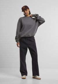 Grå oversized sweatshirt med faldende skuldre, parret med sorte breddebenede bukser med guld accenter og cremefarvede sneakers. En maroon kasket tilføjer kontrast.
