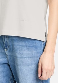 T-shirt i bomuld i lys grå med rundet nederkant, sideslids og subtil tekstdetalje nær bunden. Sammen med blå denimjeans.