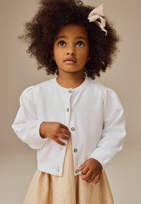 Cardigan blanc en tricot avec des manches bouffantes, dotée d'un col rond et de boutons en argent, associé à une jupe légère dorée.