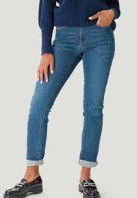 Jean en denim slim fit bleu moyen avec des ourlets retroussés, présentant un design classique à cinq poches et des coutures visibles. Porté avec des chaussures noires.