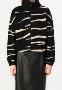 Osoba ubrana w czarno-białą w paski puszystą kurtkę z wysokim kołnierzem oraz czarną skórzaną spódnicę, z rękami swobodnie opuszczonymi wzdłuż ciała.