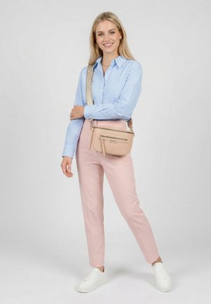 Smiling woman stands wearing light blue checkered shirt, pink pants, white sneakers, and beige crossbody bag with patterned strap on gray background.