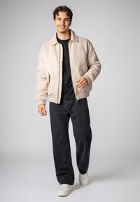 Cremfarbener Bomberjacke mit gerippten Bündchen und Kragen, kombiniert mit schwarzen Jeans und weißen Turnschuhen. Glatte Textur mit Reißverschluss.