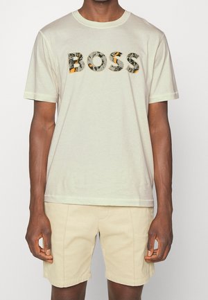 Mężczyzna w białej koszulce z krótkim rękawem, z napisem "BOSS" w czarno-pomarańczowych literach, w połączeniu z beżowymi szortami.