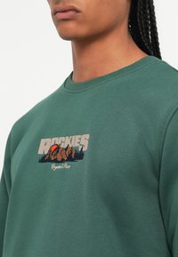 Groene sweatshirt met een ronde halslijn. Aan de voorkant is er een geborduurd ontwerp met bergen, een zon en de tekst "ROCKIES." Zachte textuur.